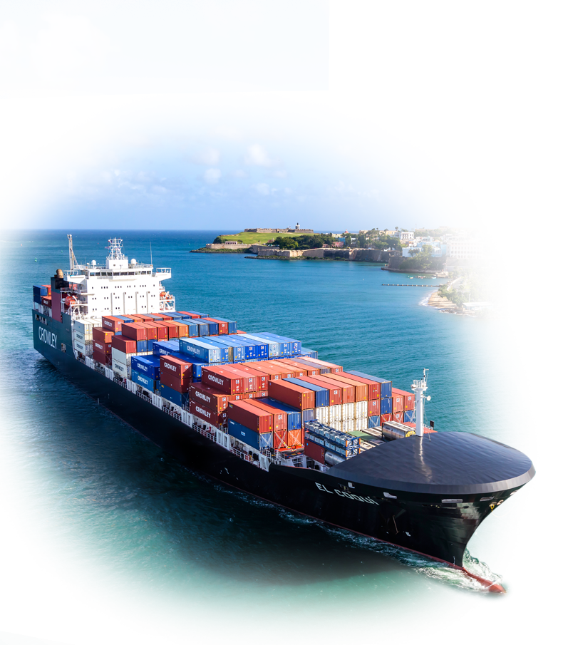 A large cargo ship loaded with colorful shipping containers sailing through a body of water, with a coastal area and buildings visible in the background under a partly cloudy sky.