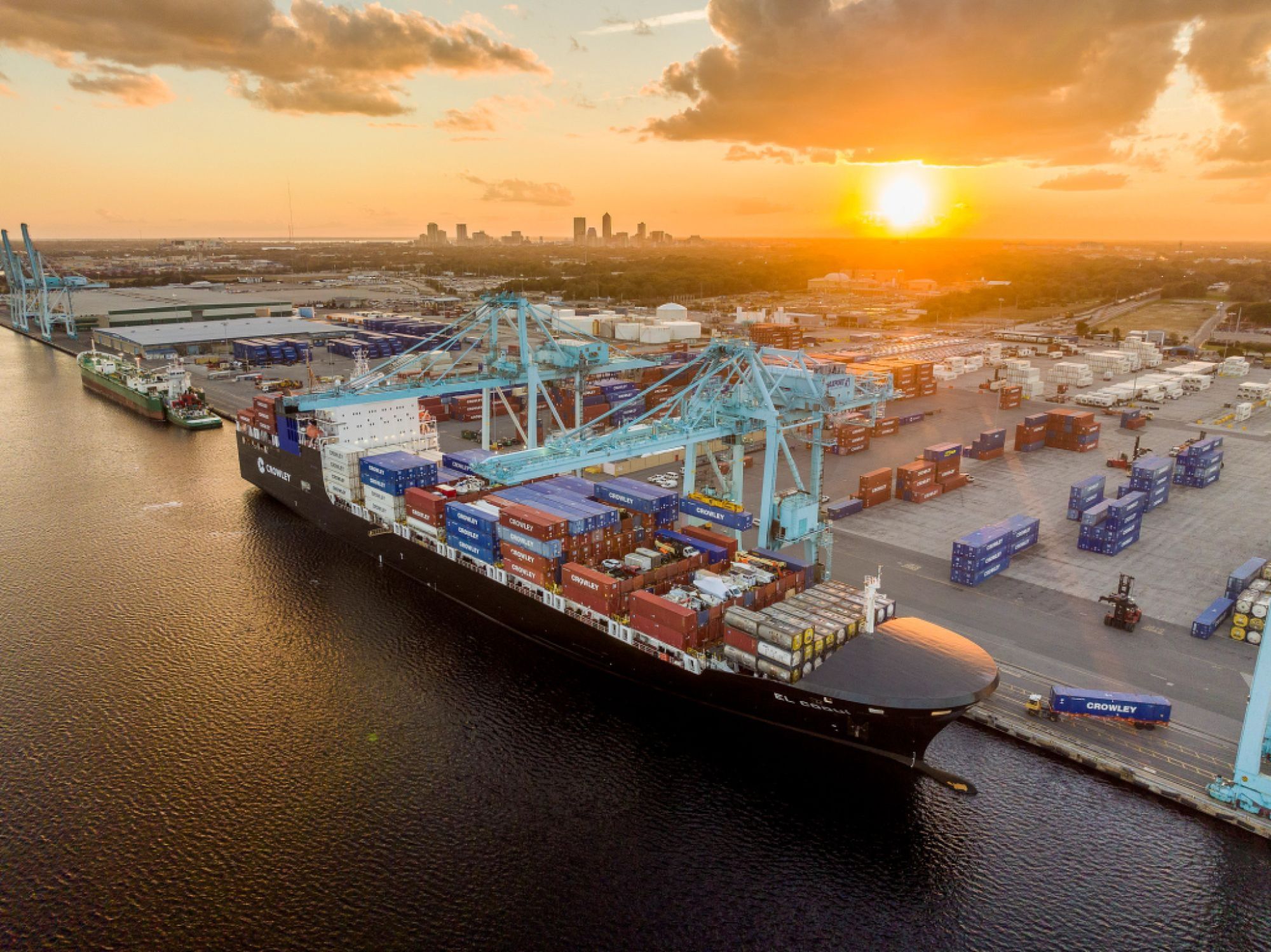 A large cargo ship docked at a busy port during sunset, with numerous shipping containers and cranes visible. The sky is partly cloudy with the sun setting in the background, casting a warm glow over the scene.