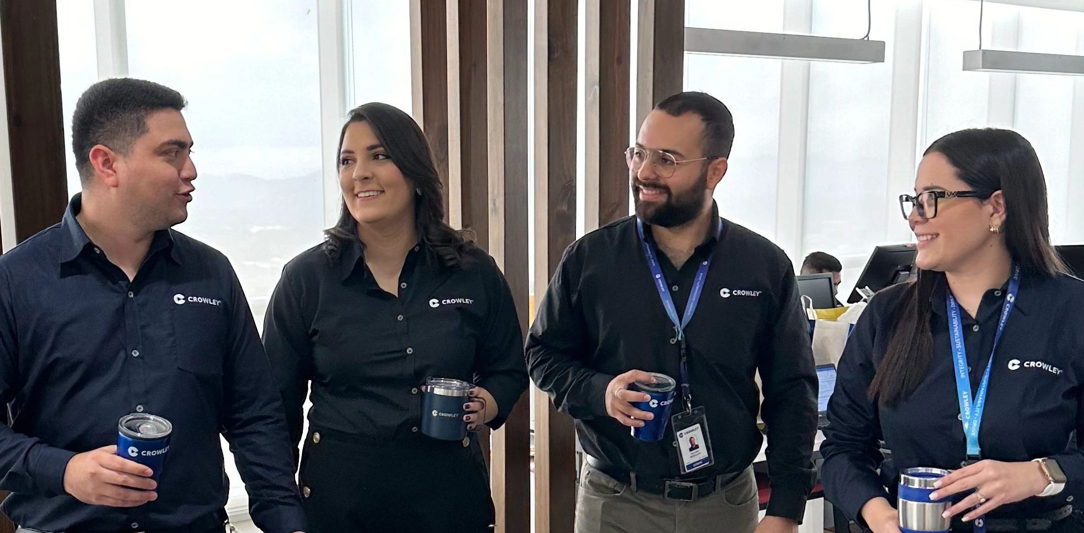 Four individuals wearing matching dark shirts with company logos stand together in an office setting, each holding a branded tumbler. They have ID badges on lanyards around their necks. The background shows modern office furniture, large windows with natural light, and vertical wooden panels.