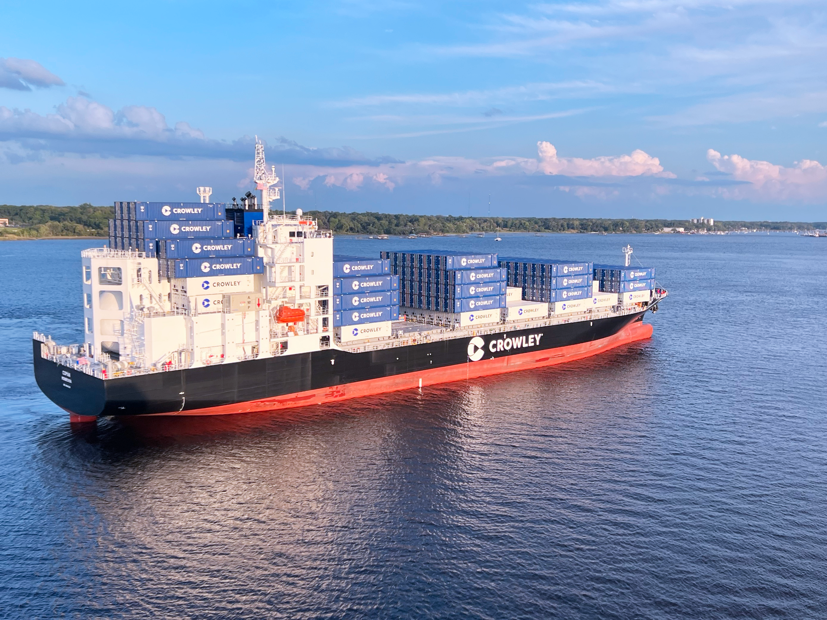 A large Crowley cargo ship named 'Copán' is sailing on a calm body of water under a partly cloudy sky. The ship has a black hull with a red bottom and is loaded with numerous blue and white shipping containers stacked neatly on its deck. The shoreline with green trees is visible in the background.