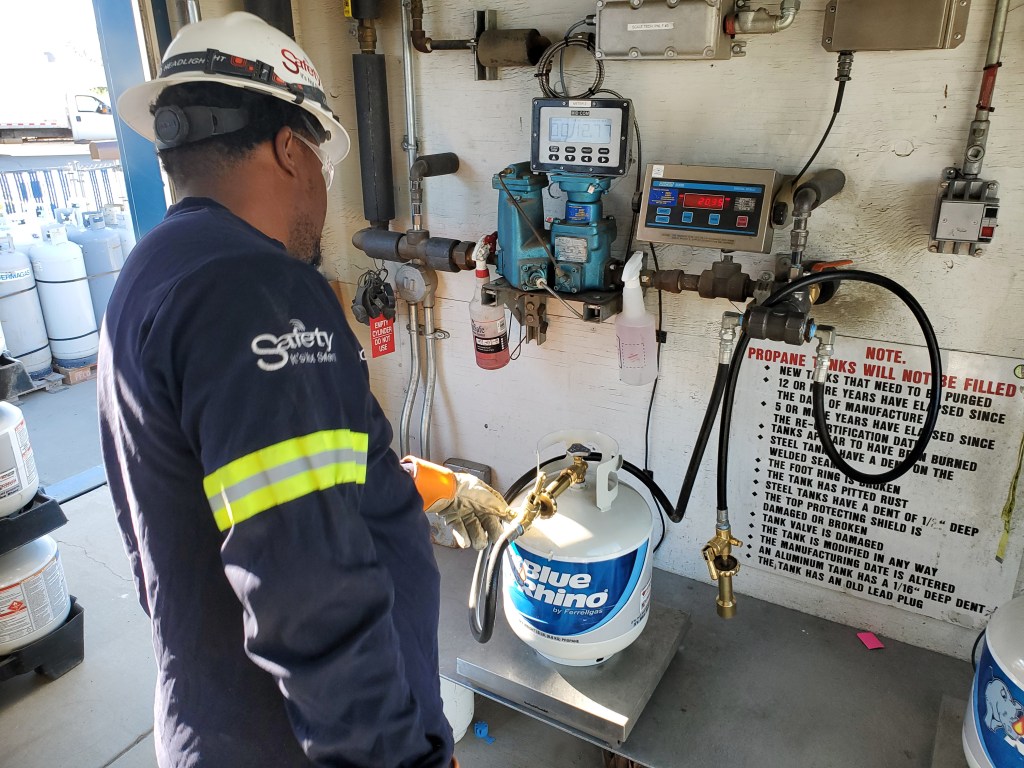 A worker in safety gear refills a Blue Rhino propane tank at a filling station. The worker wears an orange glove, white hard hat, and a navy blue shirt labeled 'Safety' on the back. The station features gauges, hoses, control panels, and a prominently displayed safety instruction sign regarding propane tank handling.