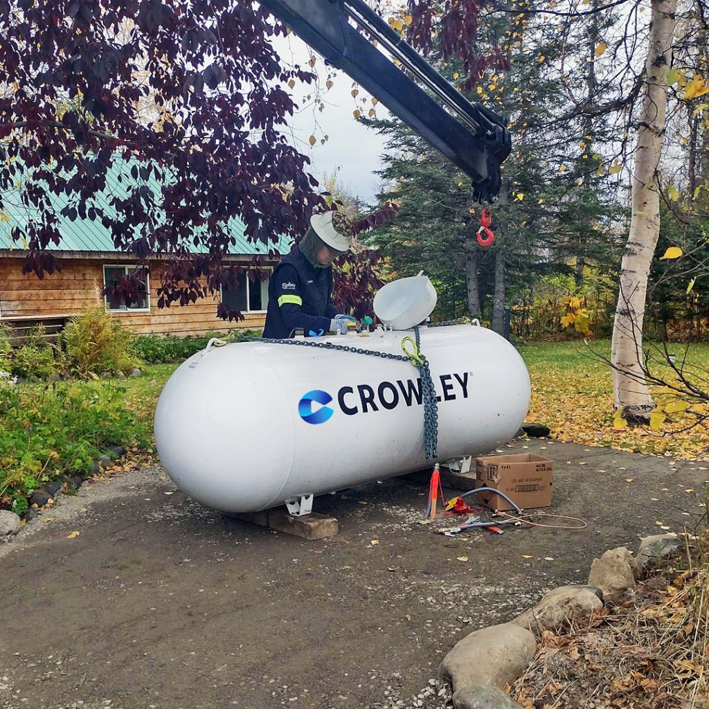 A person works on a large white tank labeled 'CROWLEY' in an outdoor area. The tank is secured with chains and positioned beneath a crane hook. The background features trees with autumn-colored leaves, a wooden building with a green roof, and various tools and equipment scattered on the ground.