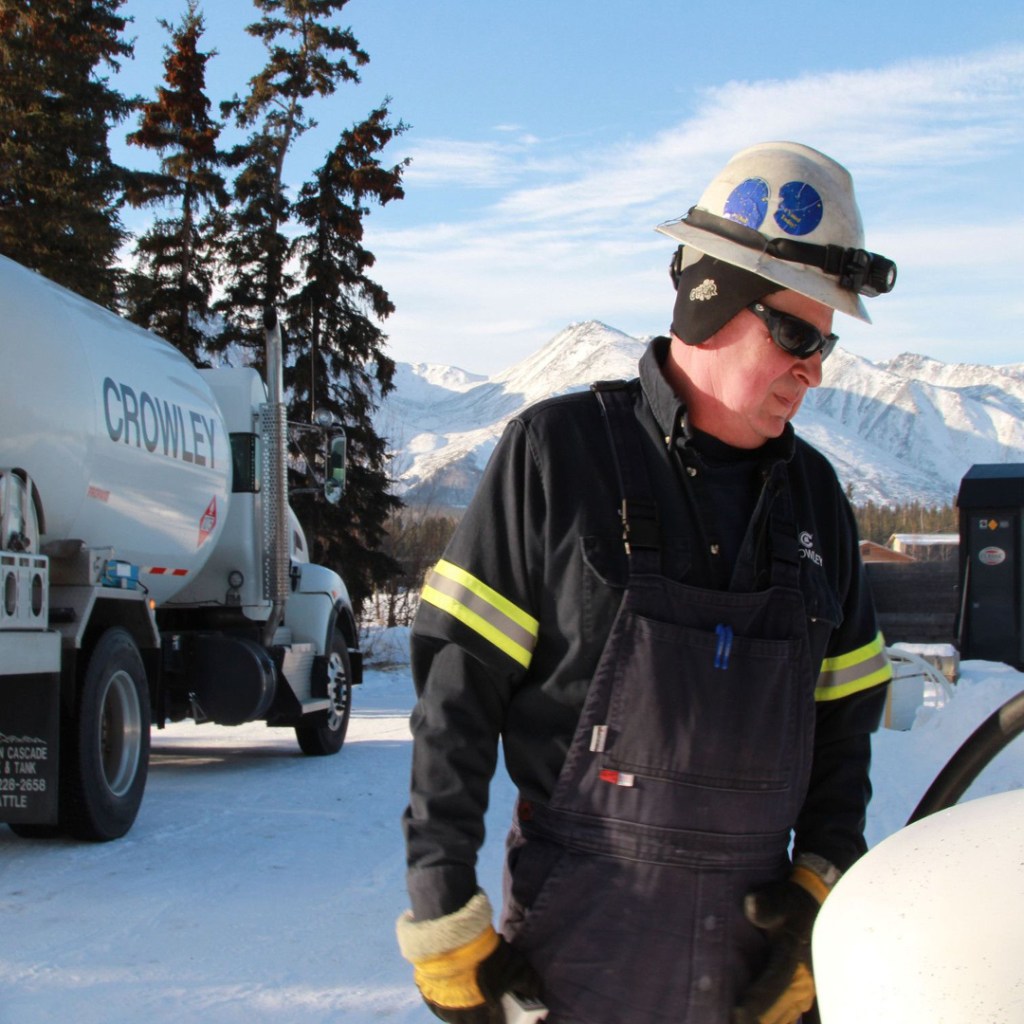 A person in winter work gear, including a hard hat and gloves, stands outdoors in a snowy environment. Behind them is a large white truck labeled 'CROWLEY,' with snow-covered mountains and trees in the background.