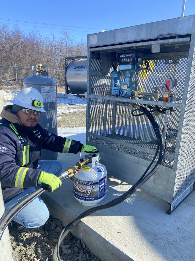 A person in safety gear, including a hard hat and gloves, kneels on the ground while working with industrial equipment outdoors. They are handling a hose connected to a large container. The setup includes gauges, valves, and multiple hoses.