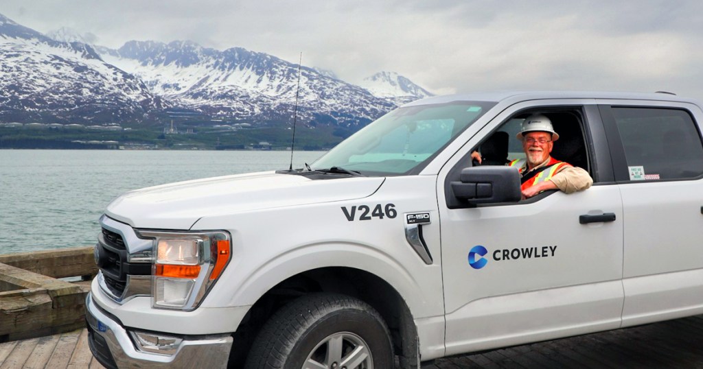 A white Ford F-150 truck labeled 'V246' on the hood and 'Crowley' on the door is parked near a body of water in Alaska, with snow-capped mountains in the background. A person wearing an orange safety vest is seated inside the truck, resting one arm on the window frame.