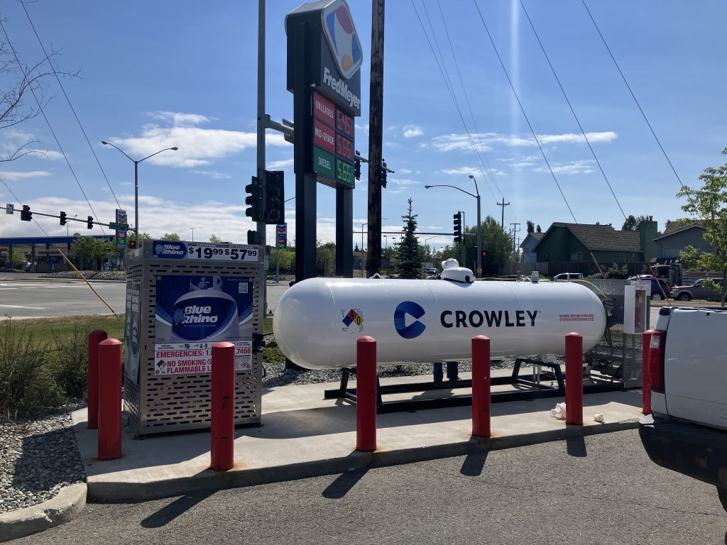 A propane tank labeled 'CROWLEY' is positioned at a gas station, surrounded by red protective bollards. In the background, a fuel price sign displays rates for unleaded and diesel fuel.