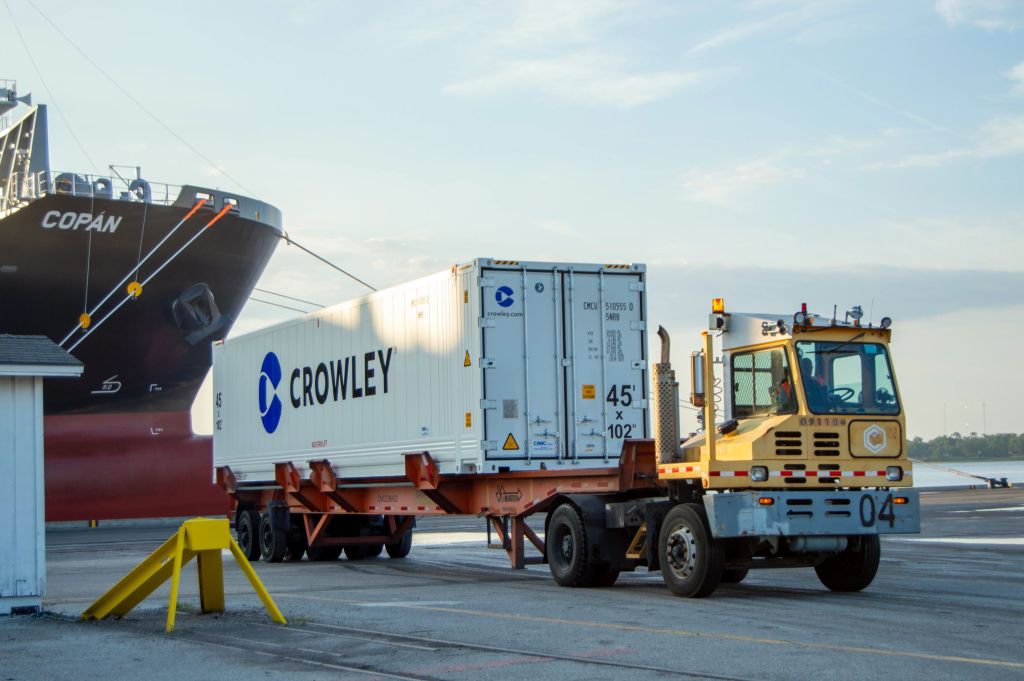 Contenedor de Crowley siendo transportado en camión en puerto, parte del proceso logístico de envío.