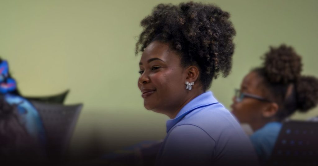 Young woman in a blue polo listening in a classroom, with another student in the background