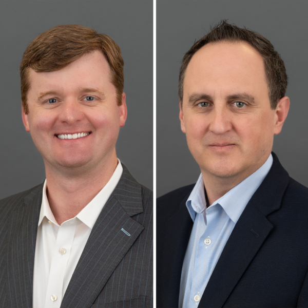 Two individuals wearing business attire against a plain gray background. The person on the left is dressed in a dark pinstripe suit with a white collared shirt, while the person on the right is wearing a dark solid-colored suit jacket over a light blue collared shirt.